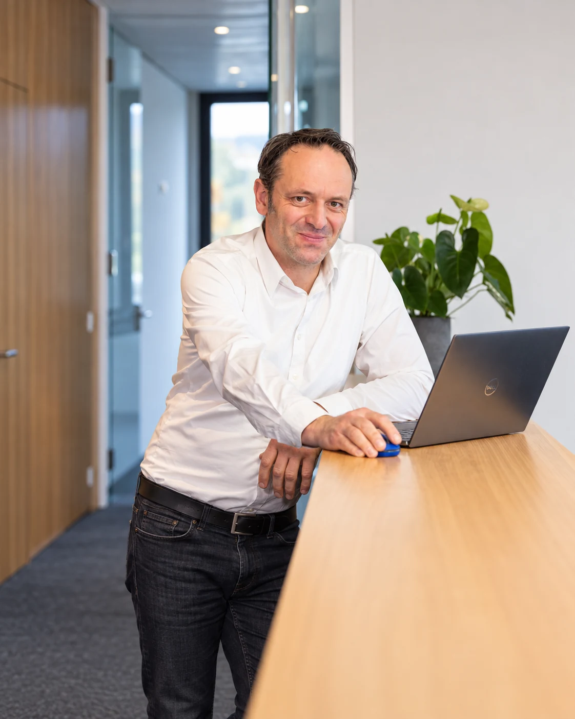 Freundlich lächelnder Mann im weißen Hemd bei der Arbeit an einem Laptop an einem Stehtresen im Büroflur