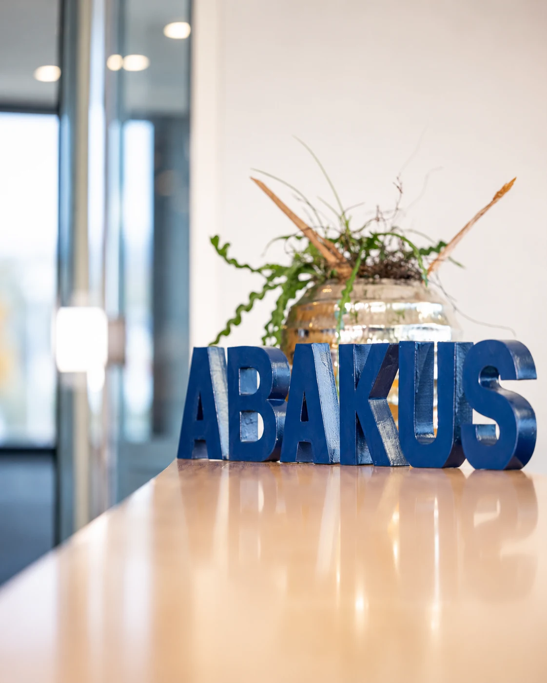 Blauer Schriftzug ABAKUS aus einzelnen Buchstaben auf einem hellen Holztresen im Büro, dahinter eine Pflanze