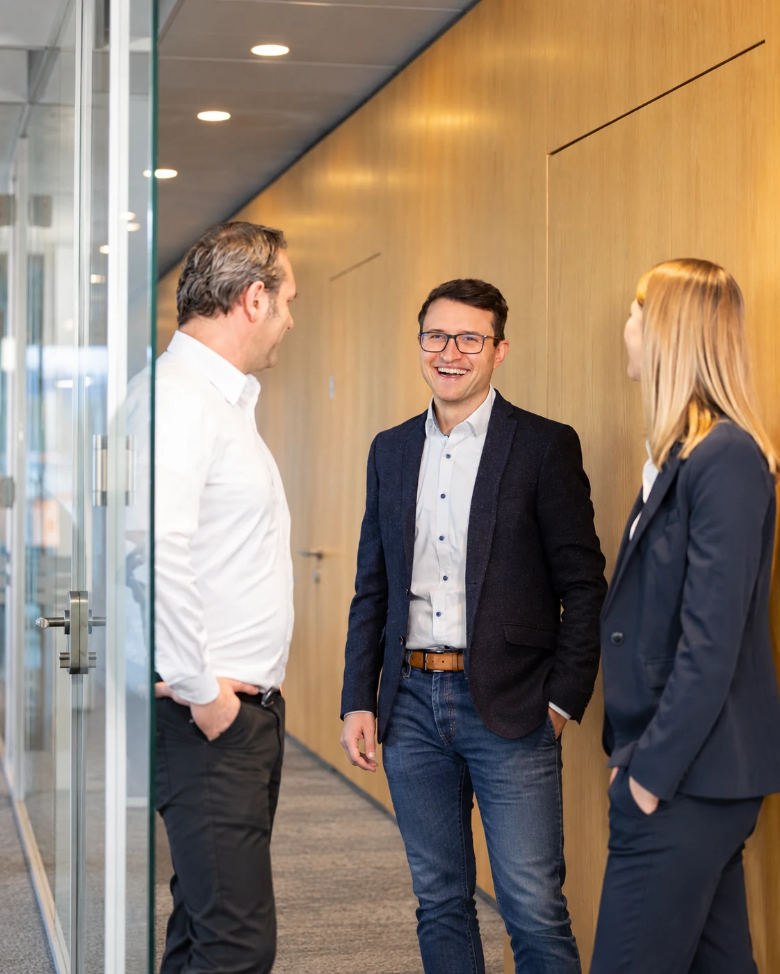 Drei Personen im Gespräch in einem Büroflur mit Glasfront und Holzwand